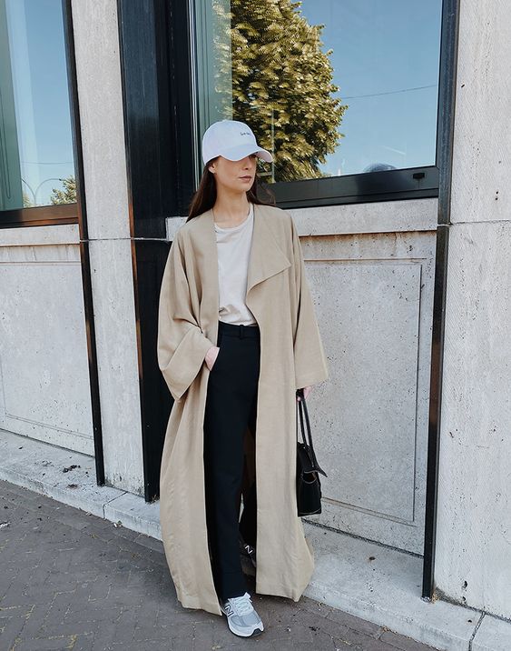a tan t-shirt, black trousers, a beige maxi trench, grey trainers, a white cap and a black tote for an everyday look