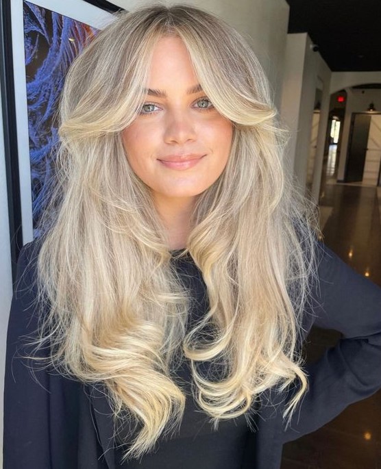 a long blonde butterfly haircut with a darker root, a money piece, a lot of volume and waves is chic and bold