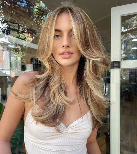 a lovely butterfly haircut on bronde hair, with blonde highlights and money piece, with feathered layers and a bit of waves is very cic