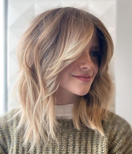 a light brown layered long bob with blonde balayage, a money piece done with an ombre effect and a lot of volume