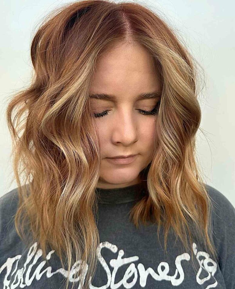 a medium length hairstyle in a ginger shade and golden blonde highlights, with  choppy layers and waves that feature texture