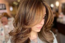 a chic brunette medium length butterfly haircut with honey balayage and curtain bangs, a lot of volume and waved ends