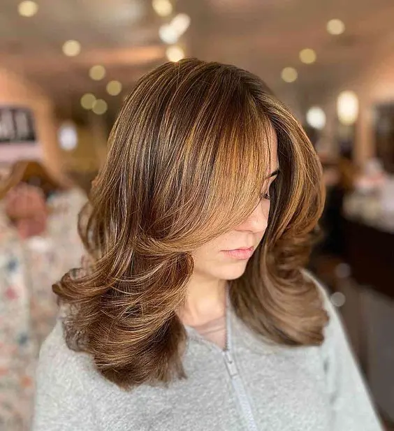 a chic brunette medium length butterfly haircut with honey balayage and curtain bangs, a lot of volume and waved ends