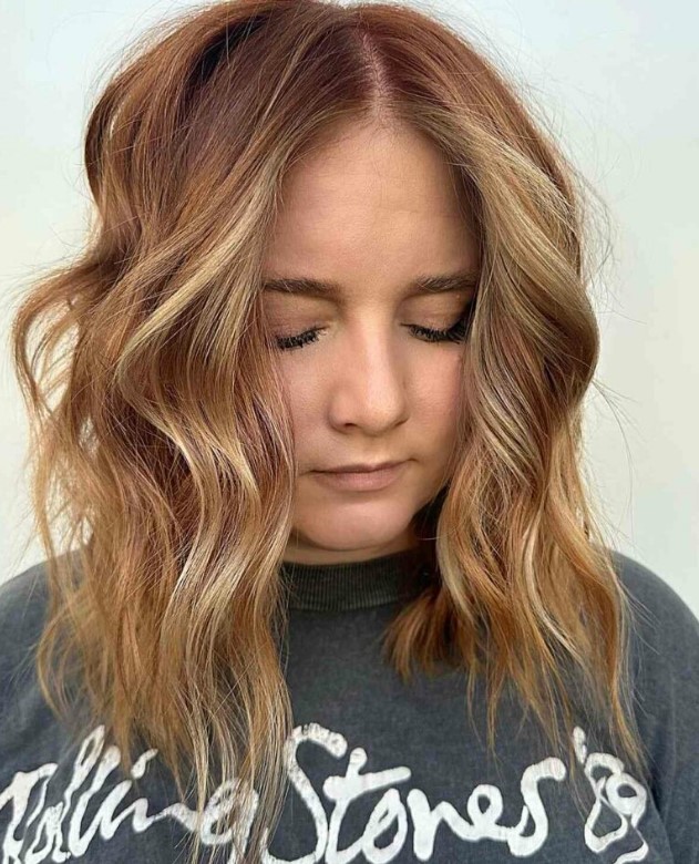 a medium length hairstyle in a ginger shade and golden blonde highlights, with choppy layers and waves that feature texture