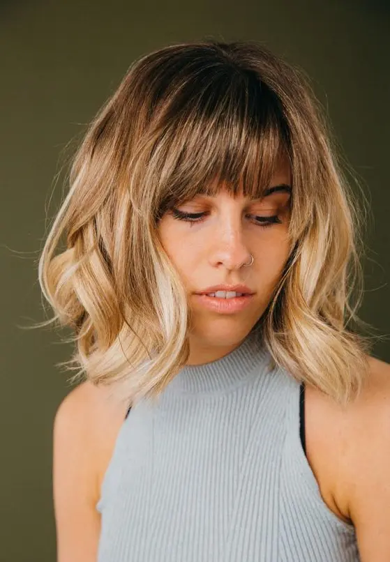 Medium length blonde hair with a darker root and waves plus wispy bangs for an effortlessly chic and cool look