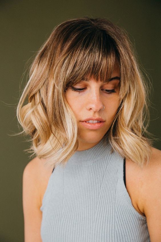 medium-length blonde hair with a darker root and waves plus wispy bangs for an effortlessly chic and cool look