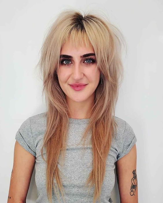 A long blonde wolf haircut with wispy bangs and a darker root looks 70s inspired and very rock style