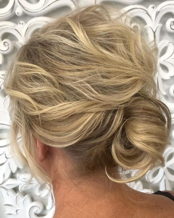 A low messy chignon with a wavy and messy top, on medium hair and with face framing locks for a mother of the bride