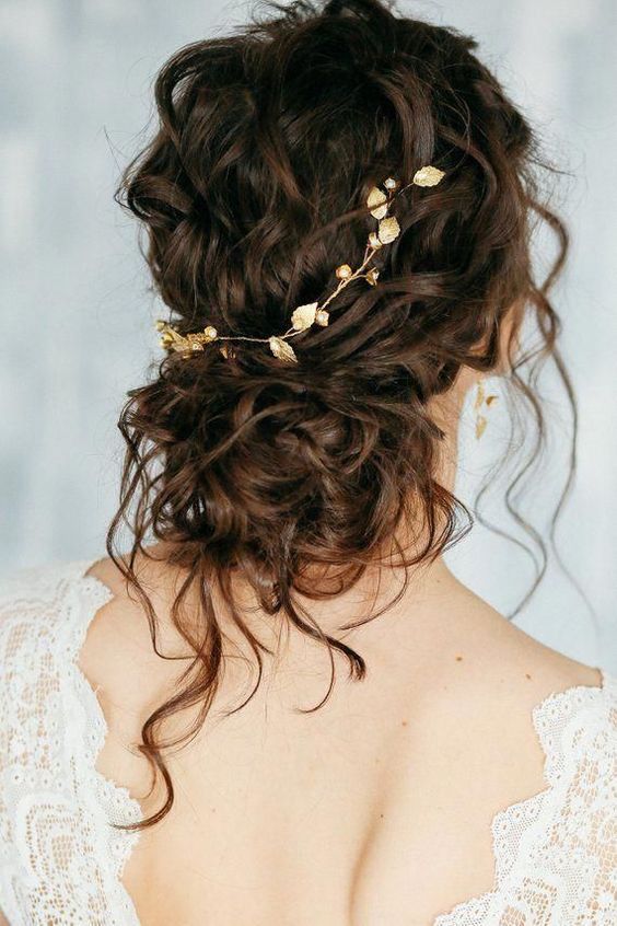 a messy curly low bun with a curly top and a chic gold leaf hair vine plus some curls down is a beautiful and subtle idea