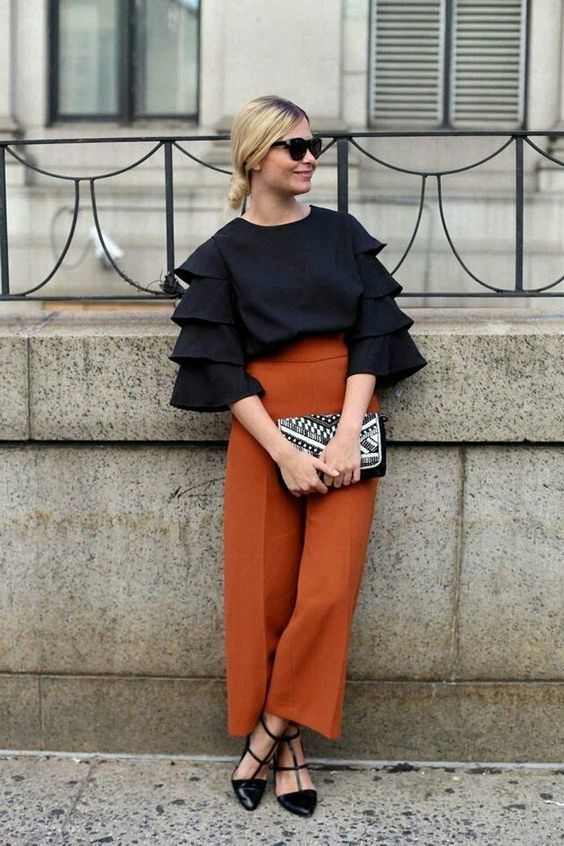 a refined fall wedding guest outfit with a black top with ruffle sleeves, rust pants and black shoes plus a black embellished bag