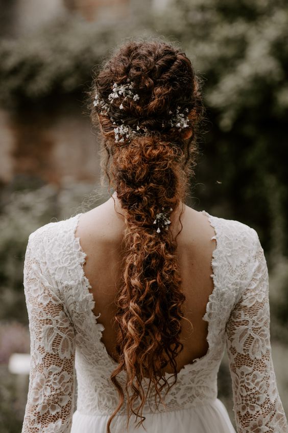 a curly low ponytail with a twisted top and a messy tail plus fresg flowers tucked in is a catchy idea for a boho bride