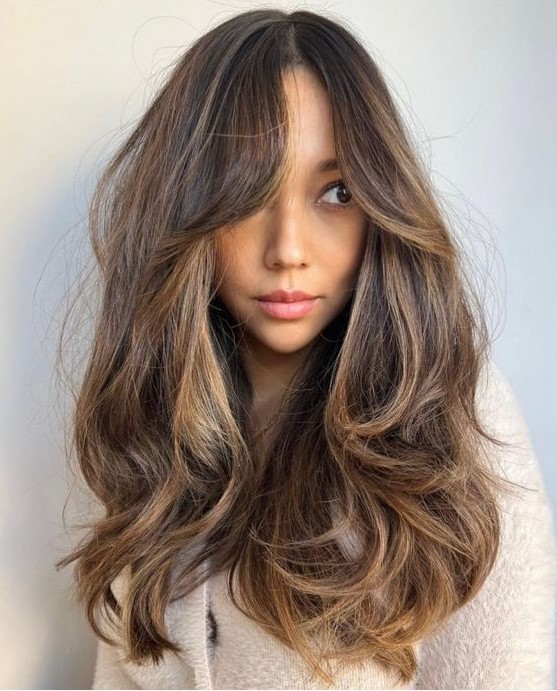 a long dark brunette hairstyle with caramel and golden blonde balayage, with bottleneck bangs and a lot of dimension and waves