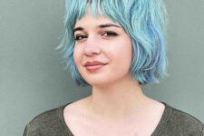 a lovely pastel blue and green shaggy messy bob with wispy bangs, a darker root and a lot of wavy volume