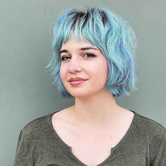 a lovely pastel blue and green shaggy messy bob with wispy bangs, a darker root and a lot of wavy volume