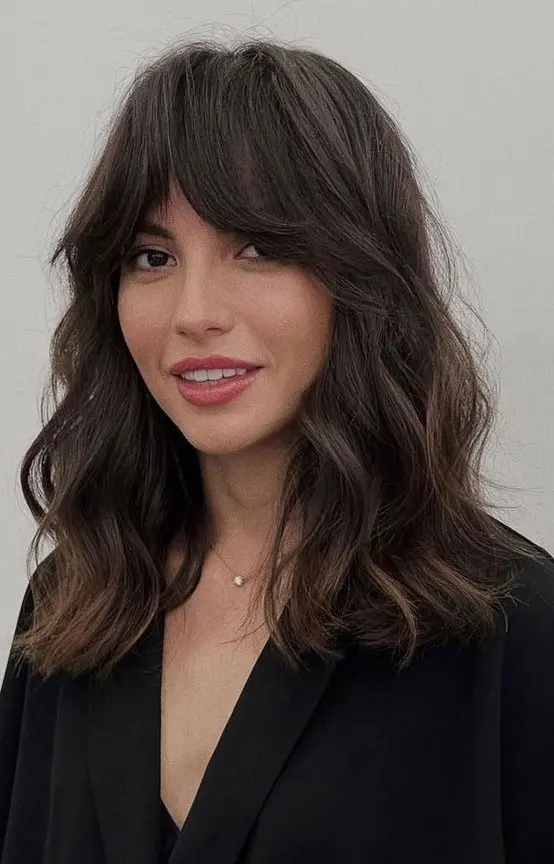 A dark brown medium length haircut with bottleneck bangs and waves plus a lot of volume looks chic and catchy