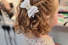 an elegant flower girl half updo with a briaded halo on top and waves down plus a white pearly bow is chic