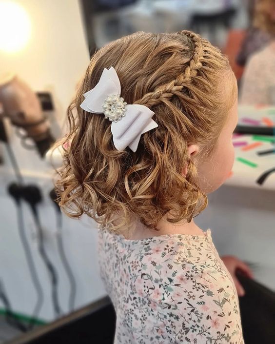 an elegant flower girl half updo with a briaded halo on top and waves down plus a white pearly bow is chic