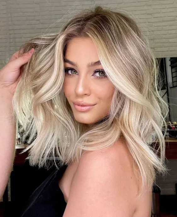 Medium length blonde hair with a shadow root and brighter balayage plus waves and volume is chic