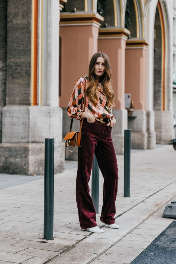 a bold and cool wedding guest look with a colorful printed top, burgundy velvet pants, white shoes and an amber bag