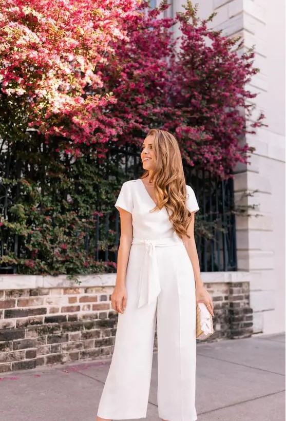 A casual white jumpsuit with a V neckline, a sash, short sleeves and a neutral bag for a spring or summer bridal shower