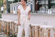 a casual white jumpsuit with a sash with a bow on the waist, nude shoes, statement earrings and a white clutch