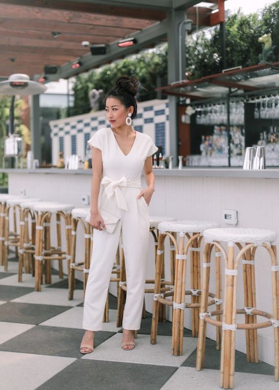 a casual white jumpsuit with a sash with a bow on the waist, nude shoes, statement earrings and a white clutch
