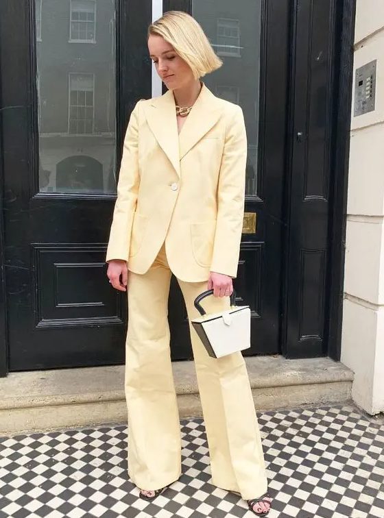 A light yellow pantsuit with flare pants and no top underneath, a two tone bag, strappy shoes and a chunky chain necklace