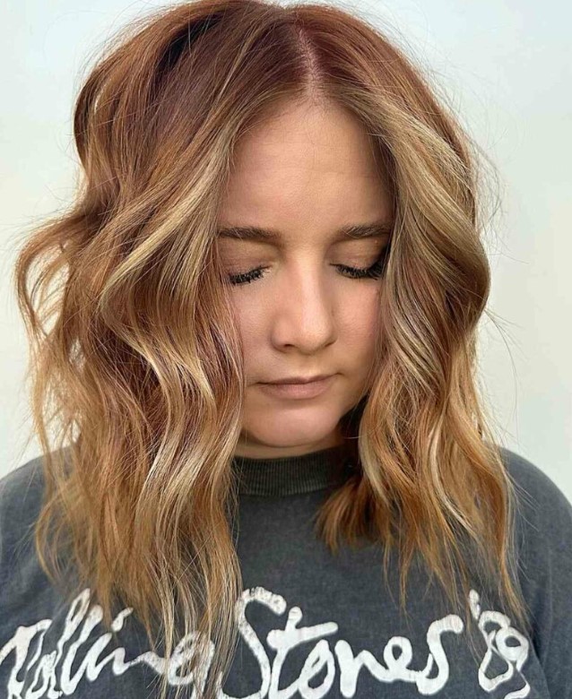 a medium length hairstyle in a ginger shade and golden blonde highlights, with choppy layers and waves that feature texture
