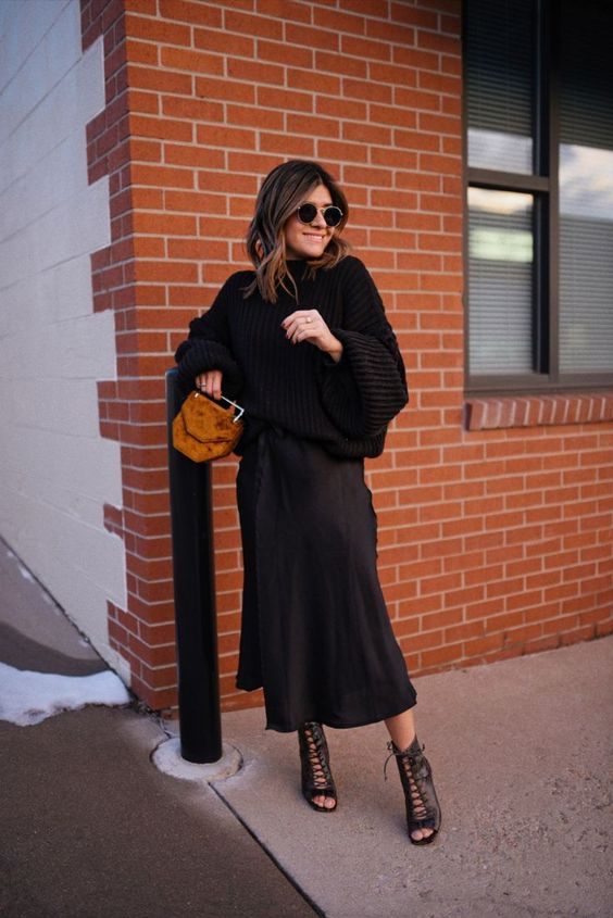 a stylish winter wedding guest outfit with a black oversized sweater, a black satin midi skirt, black lace up shoes and a brown bag