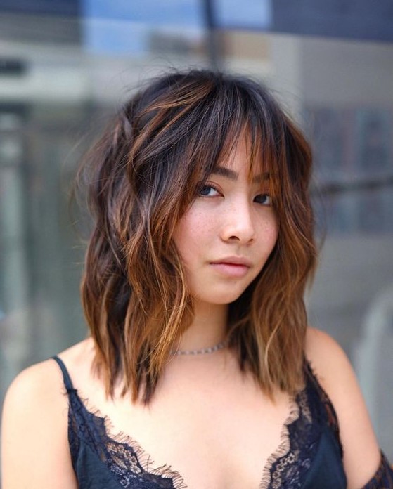 Medium length shaggy hair in dark shades and with caramel balayage, with wispy bangs to accent the eyes even more