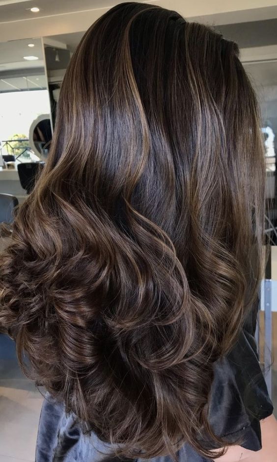 very long and extra volumetric dark brownn hair with golden blonde balayage that brings dimension and curls on the ends