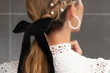 an embellished low ponytail with a braid on one side styled with pearls, rhinestones and a black velvet bow
