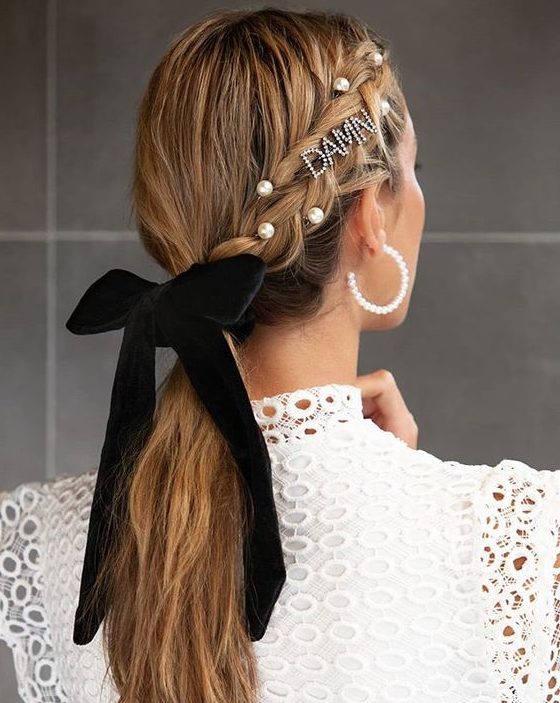 an embellished low ponytail with a braid on one side styled with pearls, rhinestones and a black velvet bow