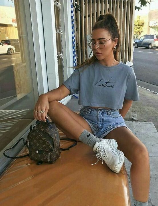A 90s inspired outfit with a cropped t shirt, blue denim shorts, white trainers and grey socks plus a brown backpack
