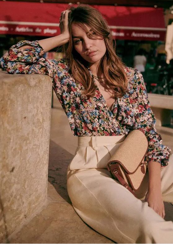 a bold floral wrap blouse with short sleeves, creamy high-waisted pants and a small bag are a cool combo for a casual summer wedding