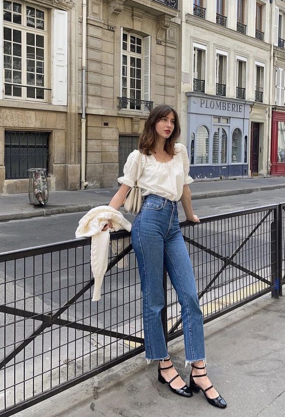 A classy Parisian chic look with a romantic ivory blouse, high waisted blue jeans, black Mary Jane shoes and a neutral cardigan