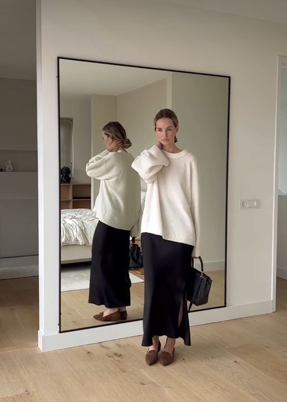 a minimalist quiet luxury outfit with a white oversized jumper, a black slip maxi skirt with a slit, brown loafers and a black bag