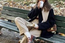 a neutral top, tan trousers, a black coat, brown loafers, white socks and a white jumper over the shoulders