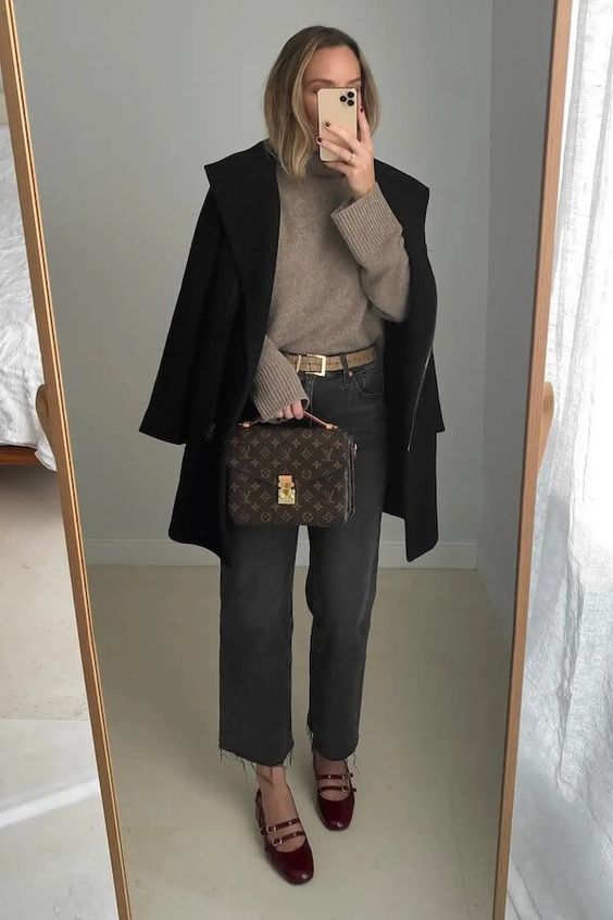 a winter or fall outfit with a greige sweater, grey jeans, burgundy Mary Jane shoes, a black coat and a brown bag