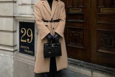 a winter quiet luxury outfit with a black turtleneck, black snakeskin print trousers, black boots, a camel midi coat, a black belt and black gloves