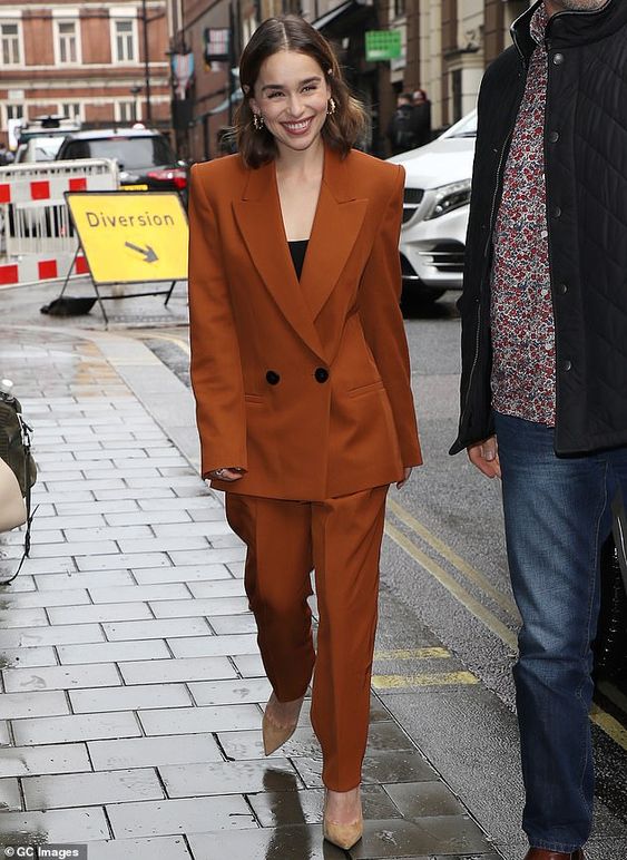Emilia Clarke wearing a terracotta power suit with accented shoulders, a black top and nude shoes looks wow
