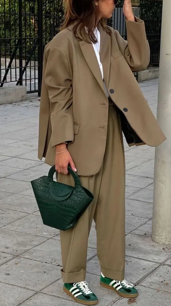 a beige oversized pantsuit, a white t-shirt, green Gazelle sneakers and a dark green bag of a catchy shape for spring