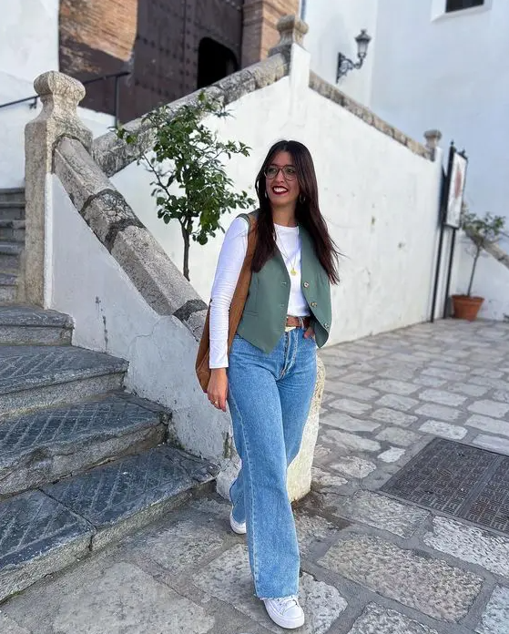 a casual outfit with a white long sleeve top, blue jeans, a green wiastcoat, white sneakers and a brown bag and belt for spring