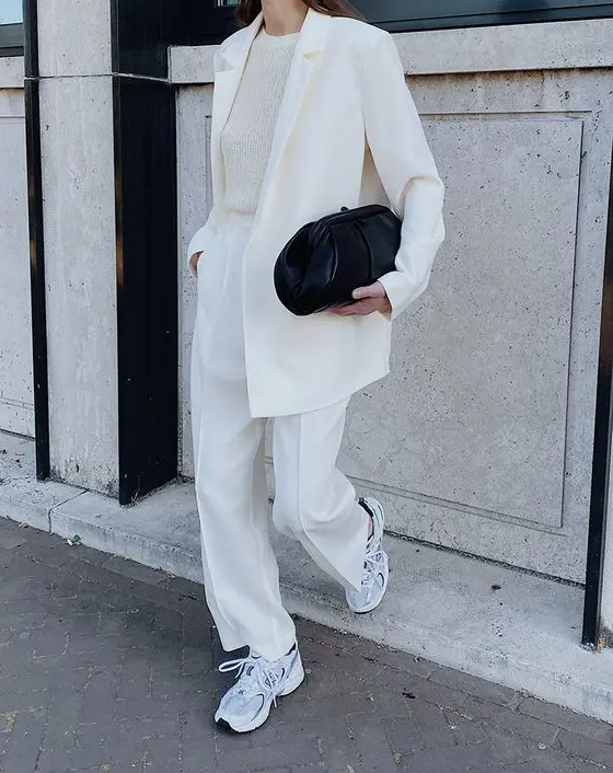 a creamy pantsuit, a neutral top, grey New Balance trainers and a black clutch are na ultimate look to rock