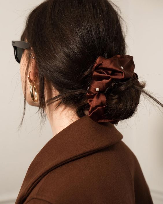 a lovely and a bit messy low bun with a shiny volumetric top and a brown scrunchie with pearls is a chic hairstyle