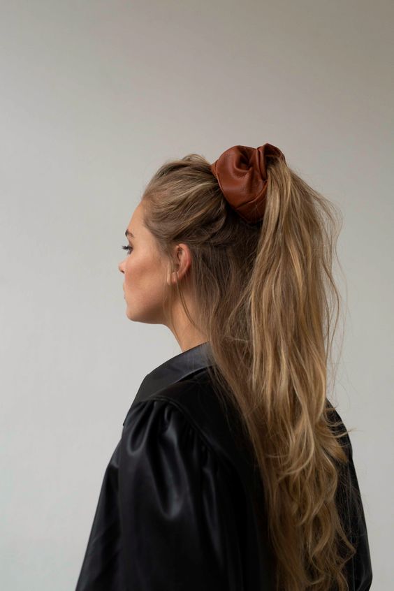 a messy half updo with a wavy high ponytail and a brown leather scrunchie as a trendy and bold accent