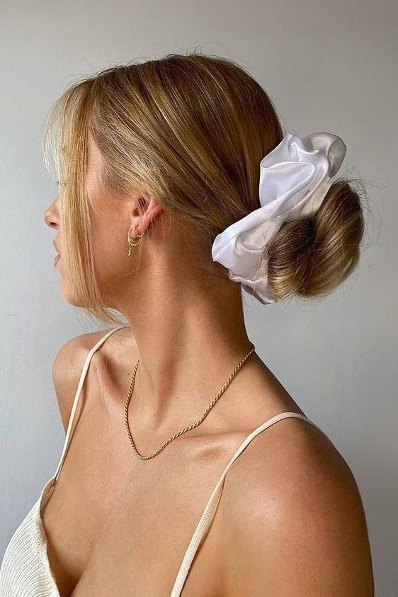 A messy wrapped low updo with a sleek top, face framing hair and a white scrunchie is a cool hairstyle
