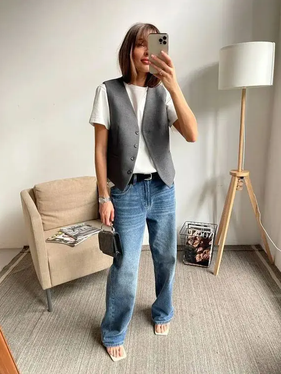 A spring outfit with a white t shirt, blue jeans, a grey waistcoat, white sandals and a small black bag