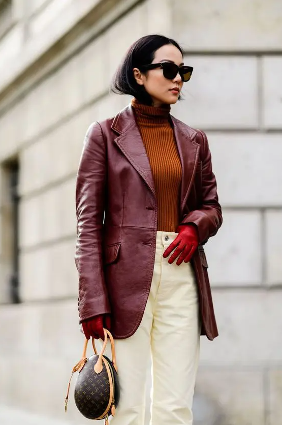 A trendy outfit with a rust colored turtleneck, white jeans, a cherry red leather blazer, red gloves and a printed bag