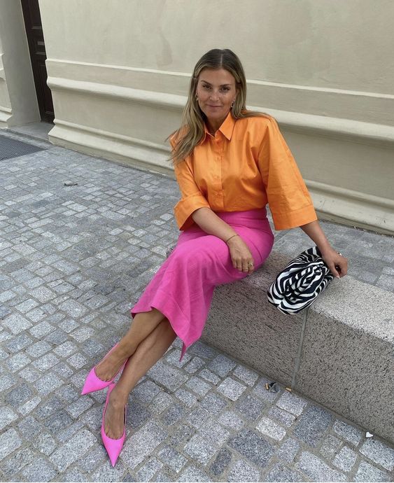 a bold spring wedding guest look with an orange shirt with wide sleeves, a hot pink midi skirt and pumps and a printed bag